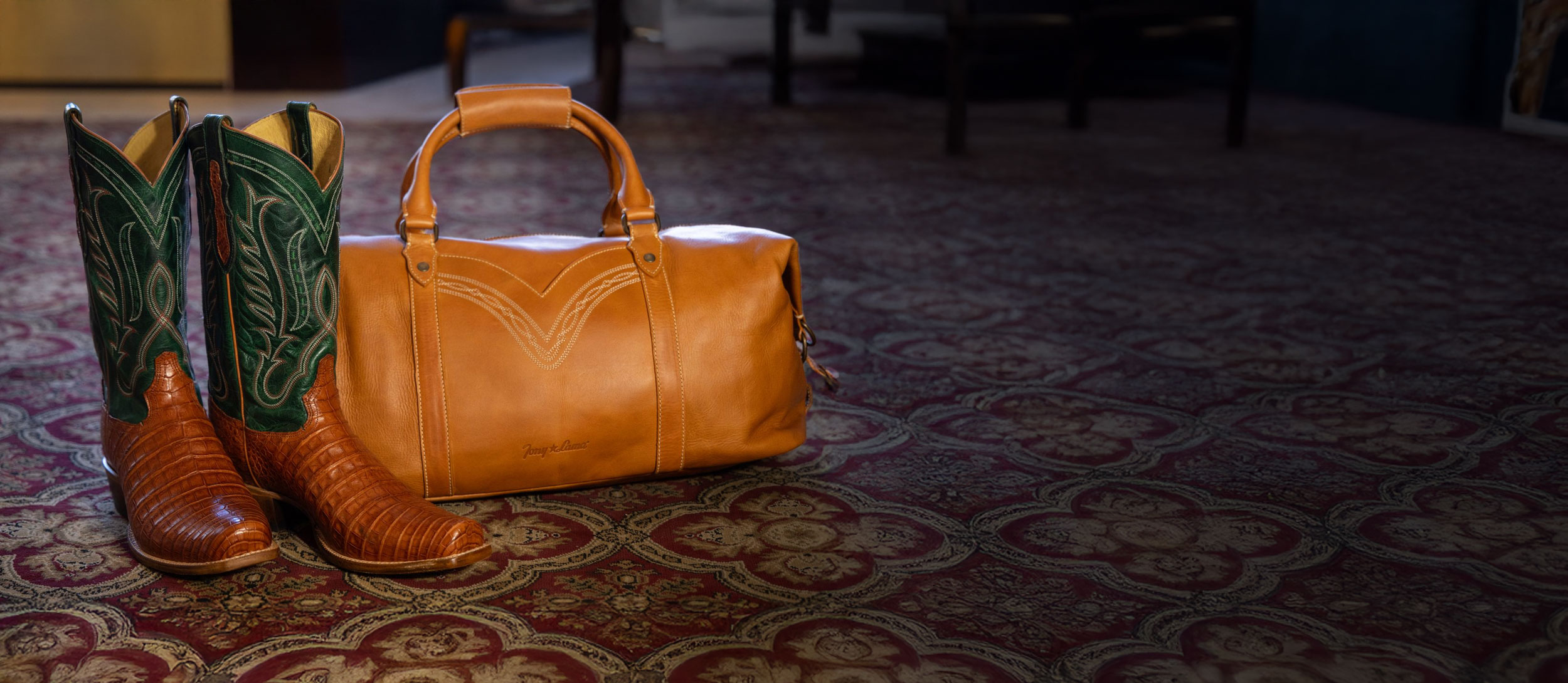 A pair of the Dinero 13" Caiman western boot in brown and green next to the Caori duffel bag in tan.